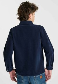 Jong persoon met kort bruin haar, gekleed in een marineblauwe corduroy overhemd en blauwe spijkerbroek, gezien van achteren tegen een effen achtergrond.