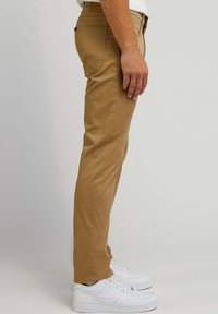 Tan chino's met een slanke pasvorm, voorzien van achterzakken en een gladde textuur, gecombineerd met witte sneakers voor contrast.