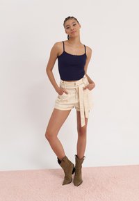 Marine ribbet tank topp, kremfargede beltehøye shorts og olivengrønne spisse ankelstøvler. Modellen poserer med hendene i lommer.