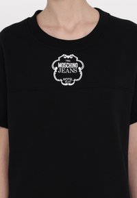 Čierne tričko s krátkymi rukávmi, na ktorom sa nachádza biela nášivka s nápisom "Moschino Jeans Hotel Milano" a dekoratívnymi prvkami. Hladká textúra.