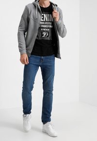 Uomo che indossa una felpa grigia con zip sopra una maglietta nera con grafiche, jeans blu e sneakers bianche, in piedi davanti a uno sfondo bianco.