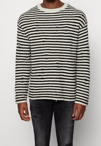 Sweter w paski w kolorze kremowym i czarnym o fakturowanym splocie, z przetarciami i okrągłym dekoltem. Połączony z ciemnymi, podartymi jeansami.