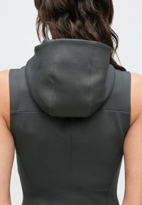 Casaco cinzento sem mangas com gola alta e capuz. O tecido tem uma textura suave e mate, e é cortado de forma impecável para um aspeto ajustado.