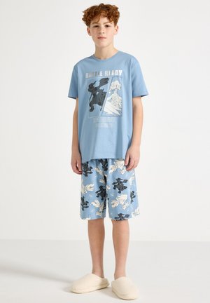 Αγόρι με κόκκινα μαλλιά που φορά μπλε μπλούζα με θέμα Pokémon και ταιριαστά σορτς, καθώς και λευκές παντόφλες, σε απλό λευκό φόντο.