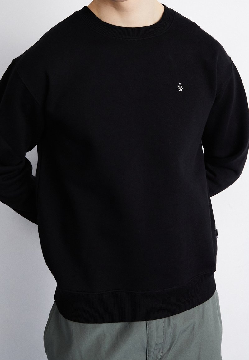Personne portant un sweat-shirt noir uni à col rond avec un petit logo blanc sur la poitrine et un pantalon gris, debout devant un fond blanc.