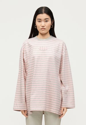 Mladá žena s dlhými tmavými vlasmi nosí oversize ružovo-šedé pruhované tričko s dlhými rukávmi, okrúhlym výstrihom a náušnicami kruhmi.