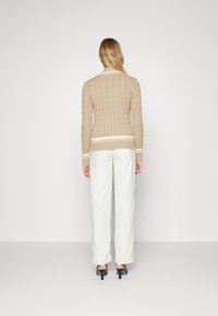 Beige kabelstickad tröja med en vit randig kant, kombinerad med vita vida byxor och gröna stiletto-klackar, sedd bakifrån.