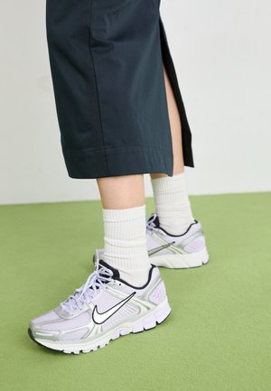 Zapatillas de running Nike en un tono lilacó con detalles plateados, logotipo negro y suela blanca. Combinadas con calcetines blancos de felpa y pantalones oscuros.