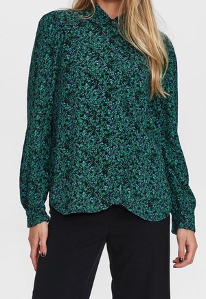 Kobieta w zielono-niebieskiej, kwiatowej bluzce z długim rękawem i czarnych spodniach, stojąca na tle jednolitego tła.