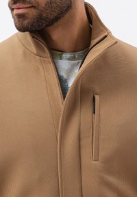 Ljusbrun zip-up jacka med strukturerad tyg, med hög krage och bröstficka. Under jackan finns en mönstrad skjorta.