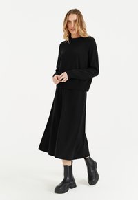 Schwarzer Strickpullover mit rundem Kragen und gerippten Bündchen, kombiniert mit einem langen schwarzen Rock. Das Modell trägt klobige schwarze Knöchelstiefel.