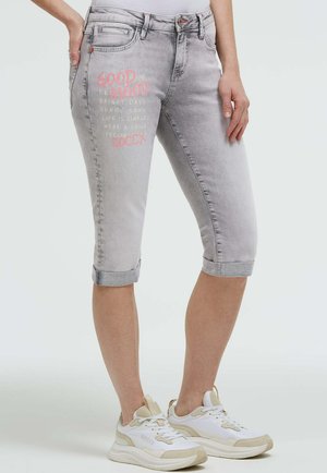 Hellgraue hochgekrempelte Jeansshorts mit pinkem und weißem Text am linken Oberschenkel, getragen mit weißen und beigen Sneakern bei einer stehenden Person.