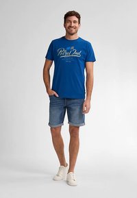 Blå bomullst-shirt med "Petrol"-grafik i ljusgul färg, kombinerad med jeansshorts och vita sneakers, stående mot en ljus bakgrund.