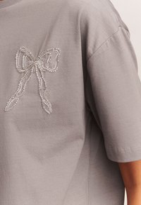 Camiseta de algodón gris con un motivo decorativo de lazo con cuentas en el pecho izquierdo, mangas cortas y un corte holgado. Textura suave.