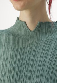 Zelená žebrovaná látka s vysokým, zaobleným výstřihem a viditelnou texturou, s jemnými horizontálními pruhy a zubatým lemem.