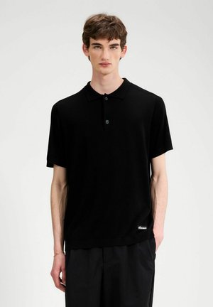 Giovane uomo con capelli castani corti, indossa una polo nera a maniche corte e pantaloni neri, in piedi contro uno sfondo chiaro e uniforme.