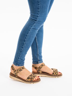 Pies con sandalias de estampado de leopardo con suelas y tiras beige, usadas con vaqueros ajustados azules sobre un fondo blanco.