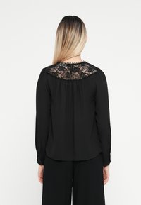 Svart langermet bluse med et blondepanel bak, strukturert stoff, og en nøkkelhullslukking i nakken med delikate trimdetaljer.