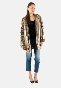 Metalen gouden pufferjack met faux-fur voering, ritsluiting, elastische manchetten en een capuchon, gecombineerd met blauwe jeans en zwarte hakken.