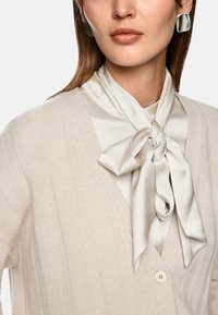 Hellbeige gerippte Strickjacke mit V-Ausschnitt und Knopfverschluss, über einer cremefarbenen Bluse und einem dekorativen Satinschal, der am Hals gebunden ist.