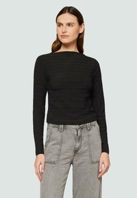 Femme avec de longs cheveux bruns portant un haut noir texturé à manches longues et un jean taille haute gris clair, regardant sur le côté.