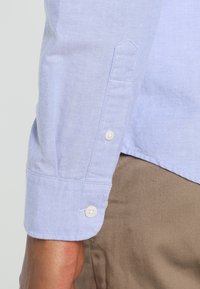 Ljusblå skjorta med knappar och en texturerad tyg. Har manschetter med knapplås och en skräddarsydd passform, i kombination med beiga byxor.