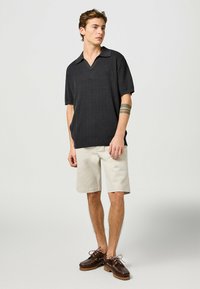 Svart stickad polotröja med korta ärmar, beige shorts och bruna mockaskor med snörning. Enkel design med avslappnad passform och v-ringning.