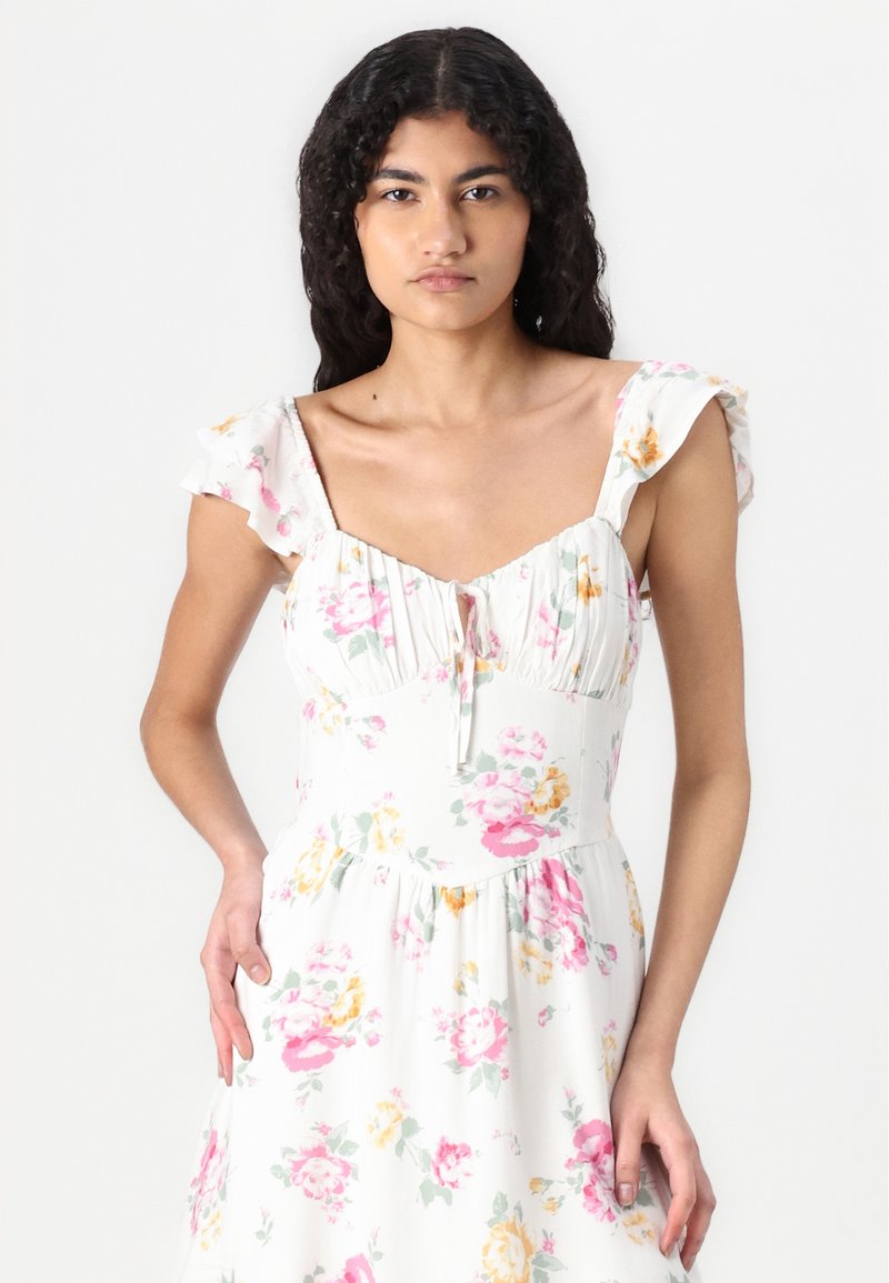 Blumenkleid mit kurzen, gerafften Ärmeln, weißem Stoff mit rosa und gelben Blumen, tailliertem Oberteil und ausgestellt Rock, mit Schleifen-Detail vorne.