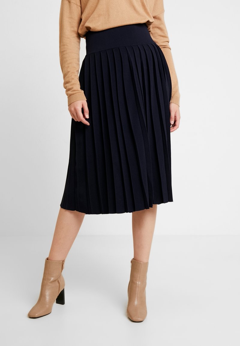 Gonna midi plissettata blu navy con una cintura larga e texture liscia, abbinata a stivali alla caviglia beige chiaro con tacco a blocco.