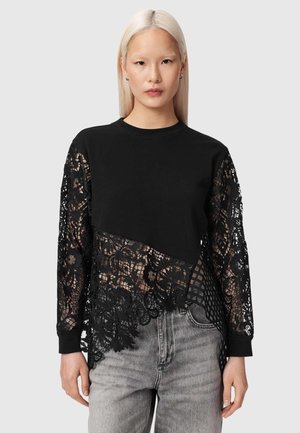 Kobieta o długich blond włosach, ubrana w czarną bluzkę z kwiatową koronkową rękawami i asymetrycznym koronkowym dołem, połączoną z jasnoszarymi dżinsami.
