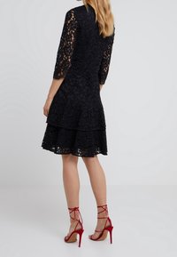 Robe en dentelle noire avec des manches trois-quarts, jupe à volants, motif floral texturé, associée à des talons hauts rouges à lacets.