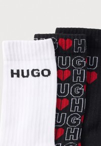 Tre paia di calzini: uno bianco con "HUGO" in nero, uno nero con "HUGH" e motivi a cuori rossi, e un altro completamente nero. Tessitura a costine.