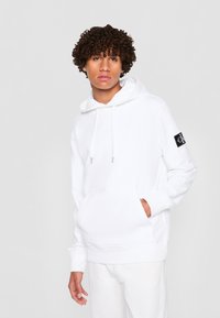 Felpa bianca con cappuccio realizzata in un misto cotone, con tasca a marsupio frontale, cappuccio con coulisse e una toppa nera con logo sulla manica.
