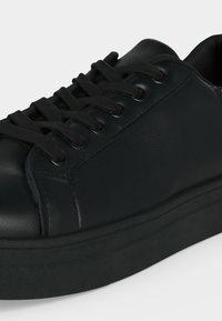 Sort lædersneaker med tyk sål og sorte snørebånd, vist i nærbillede fra siden mod en ensfarvet baggrund.