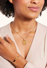 Gouden ketting met een rechthoekig hangertje en ketting, gecombineerd met een bijpassende armband, gedragen over een lichte, gestructureerde top.