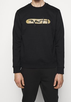 Man wearing black long-sleeve sweatshirt with beige oval and black cursive text on chest, paired with black pants, against white background.