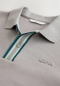 Polo gris clair avec une patte de boutonnage rayée bleue, blanche et grise et un logo CPH brodé sur la poitrine.