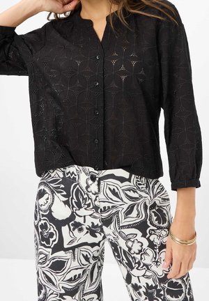 Vrouw draagt een zwarte blouse met knopen en geometrische uitsparingen, gecombineerd met hooggetailleerde zwart-witte broek met bloemenpatroon en gouden armbanden aan één pols.