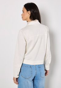 Camisa blanca corta con mangas largas y puños abotonados, con un yugo en la parte posterior y textura suave, combinada con jeans de color azul claro.