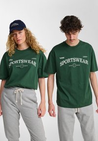 Ciemnozielone bawełniane t-shirty z białym napisem "SPORTSWEAR", w zestawieniu z szarymi spodniami dresowymi. Luźny krój; brak widocznych wzorów i elementów metalowych.