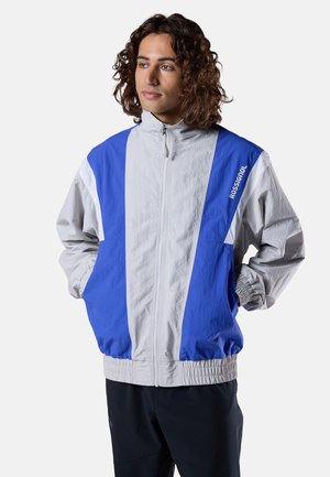 Junge Person mit lockigem Haar, die eine blau-graue Rossignol-Jacke mit Reißverschluss trägt, die Hände in den Taschen hat und seitlich schaut.