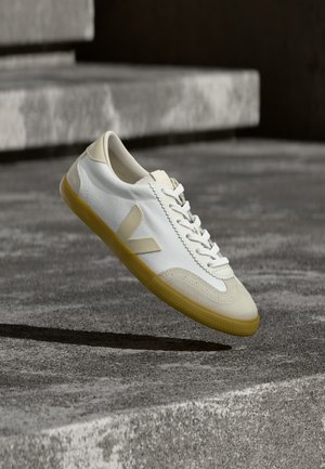 Zapatillas de lona blanca con detalles beige, punta redonda, suela de goma texturizada y el distintivo logo en forma de V en el lateral, suspendidas sobre una superficie de hormigón.
