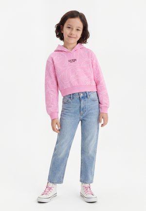 Pige iført en pink GUESS hættetrøje, lyseblå jeans og hvide sneakers med pink snørebånd mod en ensfarvet hvid baggrund.