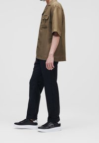 Olijfgroene shirt met korte mouwen en een borstzak, gecombineerd met zwarte broeken en zwarte sneakers met een witte zool en logo.