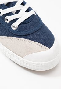 Marineblaue Leinensneaker mit beigem Wildlederakzent, weißer Gummikappe und weißen Schnürsenkeln, die durch silberne Ösen gefädelt sind. Texturierte Oberfläche.