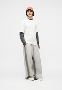 Νεαρός που φοράει κόκκινο καπέλο, λευκό κοντομάνικο πουκάμισο πάνω από μακρύ μαύρο μανίκι, φαρδιά γκρι παντελόνια και λευκά αθλητικά παπούτσια, στέκεται με τα χέρια στις τσέπες.