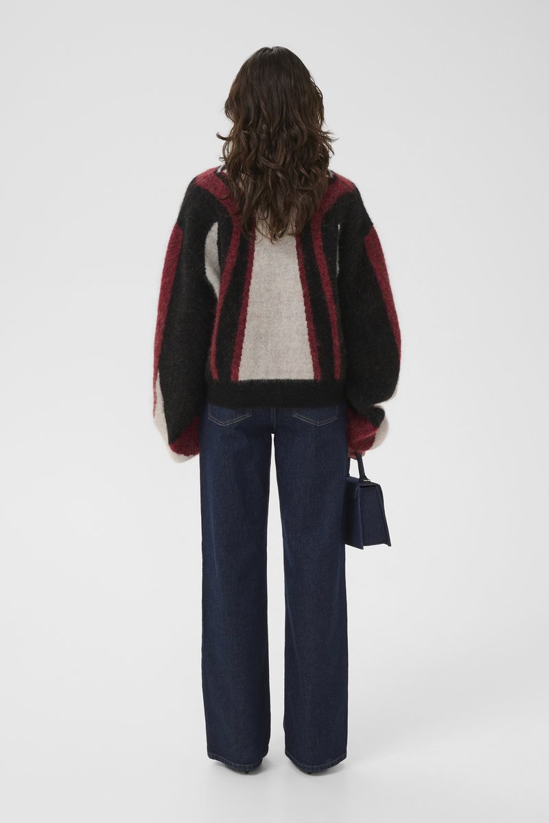 Donna con capelli castani mossi che indossa un maglione oversize nero, rosso e beige, jeans larghi blu scuro, mentre tiene una piccola borsa a tracolla blu navy.
