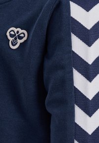 Marinblå sweatshirt med strukturerad tyg, broderad bi-logotyp och vita chevronränder längs sidan.