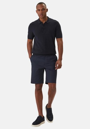 Hombre caminando con un polo negro de manga corta, pantalones cortos azul marino y zapatillas negras con suelas blancas sobre un fondo blanco.