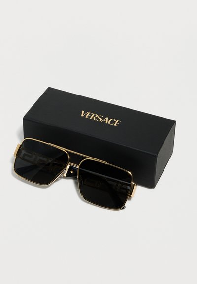 Μαύρα και χρυσά ορθογώνια γυαλιά ηλίου με μοτίβο ελληνικού κλειδιού στους φακούς, τοποθετημένα μπροστά από μια μαύρη κουτί με το λογότυπο της Versace.